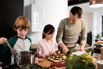 Ein Vater kocht mit seinen beiden Kindern. Die Tochter bereitet das frische Gemüse vor, der Sohn rührt im Kochtopf. Hier trifft viel virtuelles Wasser auf nur geringen realen Wasserverbrauch.