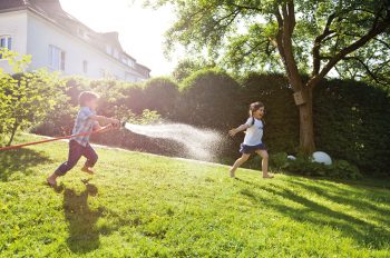 Kinder spielen im Garten. Mit großem Vergnügen spritzen sie sich mit dem Gartenschlauch nass. Hier macht Wasser sparen wirklich keinen Sinn.