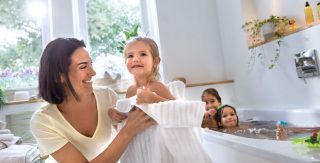 Im Hintergrund planschen zwei Kinder in der Badewanne. Im Vordergrund trocknet eine Mutter ihr Kind ab.