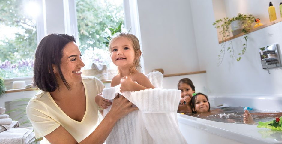 Im Hintergrund planschen zwei Kinder in der Badewanne. Im Vordergrund trocknet eine Mutter ihr Kind ab.