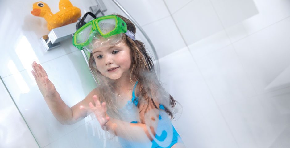 Ein kleines Mädchen, in blauem Badeanzug und mit grüner Taucherbrille, malt an dem beschlagenen Glas in der Dusche.