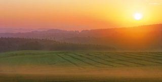 Blick über eine endlos weite Landschaft mit Wiesen und Wäldern, im Hintergrund geht leuchtend die Sonne auf.