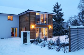 Ein Haus in winterlicher Abenddämmerung. Alles ist schneebedeckt, durch die Fenster scheint stimmungsvolles Licht in den Garten. Dort steht die Außeneinheit einer Wärmepumpe. Diese Elektroheizung versorgt das ganze Haus mit Wärme.