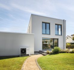 Der Blick auf die Terrasse eines neu gebauten Eigenheims. An der Rückseite der angrenzenden Garage ist die Außeneinheit einer Luft-/Wasser-Wärmepumpe aufgestellt.