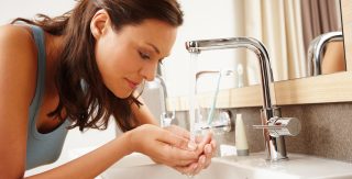 Eine Frau mit langen braunen Haaren schöpft mit beiden Händen an einem Waschtisch Wasser aus einem Wasserhahn