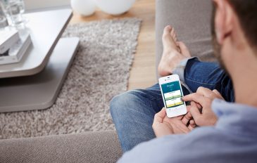 Ein Mann sitzt mit dem Smartphone in der Hand im Wohnzimmer und wählt per App seine Wunschtemperatur aus.