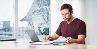 Eine Mann sitzt mit seinem Laptop und vielen Papieren, die er prüfend anschaut, an einem Tisch vor einem Fenster.