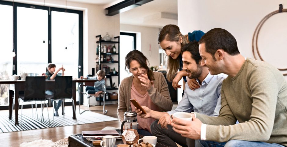 4 fröhliche junge Menschen sitzen in einem offenen Raum an einem Tisch und betrachten etwas auf einem Smartphone.