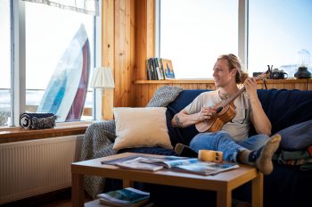 Ein Mann sitzt entspannt auf dem Sofa und spielt Gitarre; im Hintergrund sind durch die Fenster Himmel und Meer zu sehen.