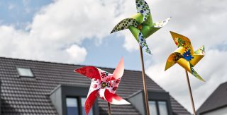 Drei bunte Kinderwindmühlen drehen sich im Wind. Dahinter ist ein Haus zu sehen.