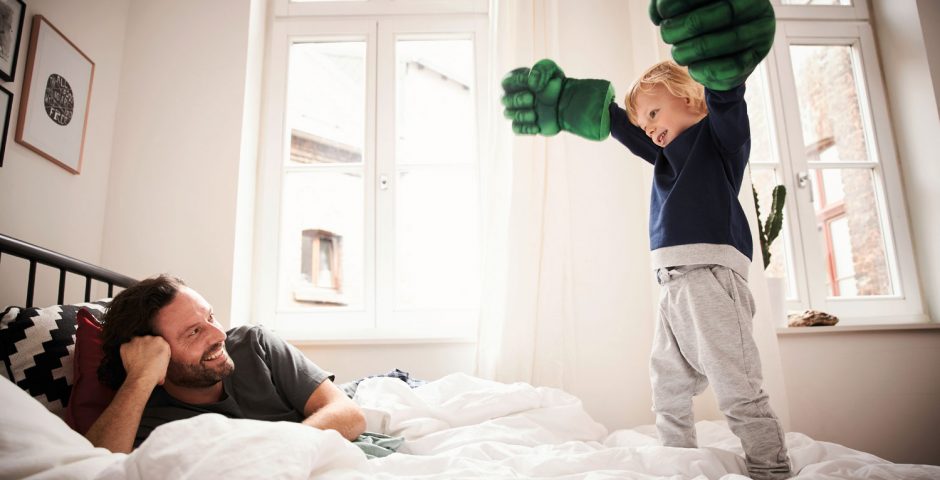 Ein Vater spielt mit seinem Sohn im Bett. Im Hintergrund sind große geschlossene Fenster zu sehen.