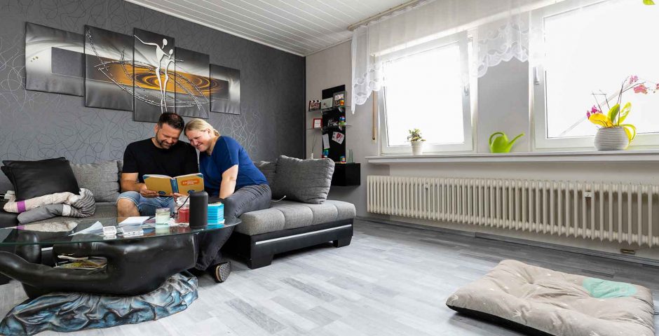 Ein Paar sitzt im Wohnzimmer gemütlich auf dem Sofa und schaut gemeinsam in ein Buch. Unter dem langen Fenster ist ein großer Heizkörper.