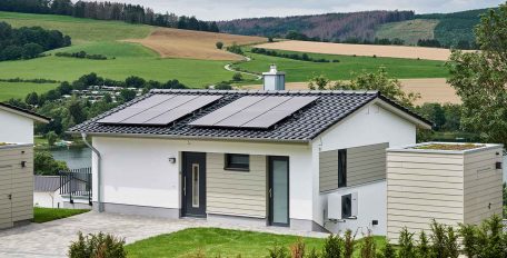 In einer grünen, sonnigen Landschaft steht ein kleines Einfamilienhaus am Hang, das mit einer Wärmepumpe beheizt wird.