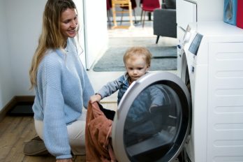 Eine Frau sitzt lächelnd mit einem kleinen Kind vor einer Waschmaschine.