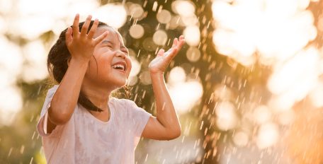 Ein Kind in einem weißen Shirt spielt lachend im Regen, die Hände neben dem Kopf erhoben.