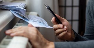 Ein Mann hält sein Handy und eine Abrechnung in der Hand und kniet vor einem Heizkörper