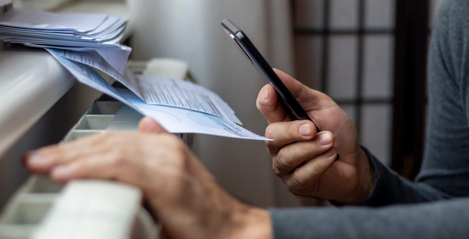 Ein Mann hält sein Handy und eine Abrechnung in der Hand und kniet vor einem Heizkörper