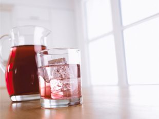 Eine Karaffe mit aromatisiertem, rötlichem Wasser steht auf dem Tisch neben einem gefüllten Glas, in dem Eiswürfel gestapelt sind.