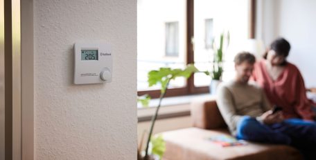 An der Wand hängt ein Raumtemperaturregler; im Hintergrund sitzt ein Paar vor dem Fenster auf einer Couch.