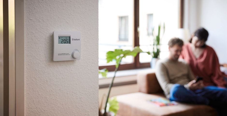 An der Wand hängt ein Raumtemperaturregler; im Hintergrund sitzt ein Paar vor dem Fenster auf einer Couch.