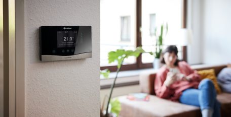 Im Vordergrund hängt ein digitaler Heizungsregler von Vaillant an einer Zimmerwand, im Hintergrund sitzt eine Frau auf einer Couch und schaut auf ihr Smartphone