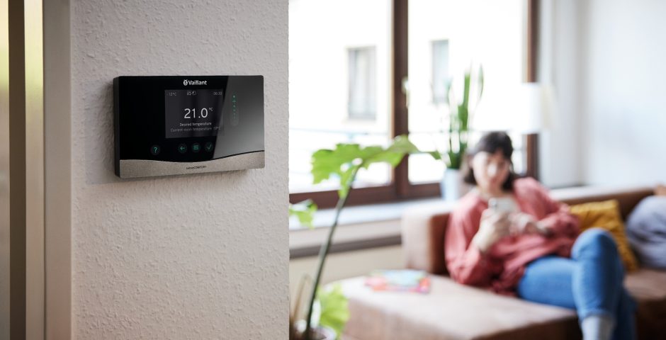 Im Vordergrund hängt ein digitaler Heizungsregler von Vaillant an einer Zimmerwand, im Hintergrund sitzt eine Frau auf einer Couch und schaut auf ihr Smartphone