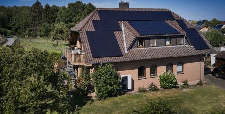 In einer grünen Landschaft steht ein verklinkertes Einfamilienhaus mit Walmdach. Darauf befindet sich eine große PV-Anlage.