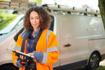 Frau in orangefarbener Arbeitsjacke mit einem Tablet in der Hand vor einem weißen Lieferwagen.