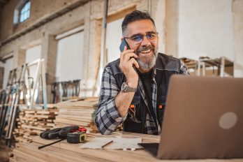 Handwerker in einer holzverarbeitenden Werkstatt telefoniert per Smartphone mit Blick auf das Laptop.