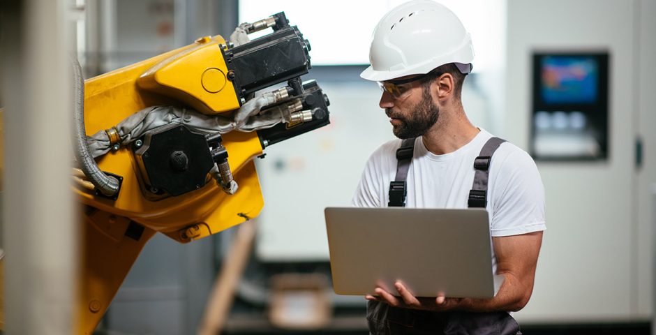 Mann in Arbeitslatzhose mit weißem Bauhelm und Laptop in der Hand schaut auf eine gelbe Maschine