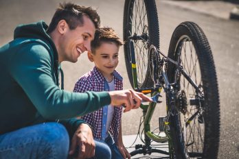 Ein Mann und ein Junge knien lächelnd vor einem Fahrrad, das auf Sattel und Lenker steht. Der Mann hält ein Werkzeug in der Hand.