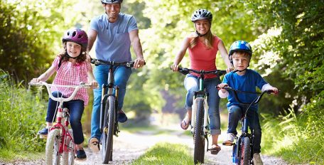 Eine Familie bestehend aus Mutter, Vater, einem Mädchen und einem Jungen fahren bei Sonnenschein über einen Waldweg.
