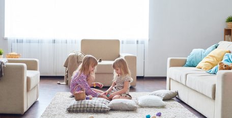 In einem sonnendurchfluteten Wohnzimmer sitzen zwei jüngere Mädchen zwischen Sofa und Sesseln auf einem weißen flauschigen Teppichboden, umgeben von hellen Kissen sind sie versunken in ihr ruhiges Spiel mit bunten Kugeln.