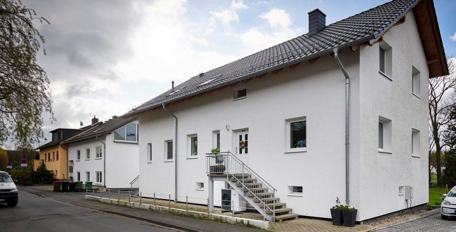 Unter der Treppe zur Haustür eines älteren Einfamilienhauses steht eine Luft/Wasser-Wärmepumpe.