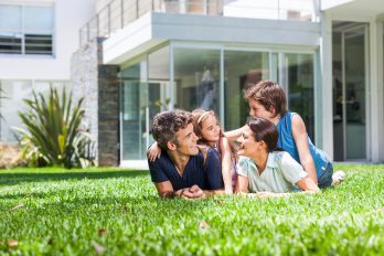 Vor einem modernen Haus liegt eine Familie bestehend aus Mutter, Vater und zwei Kindern bäuchlings auf dem Rasen, die sich glücklich lächelnd einander zuwenden.