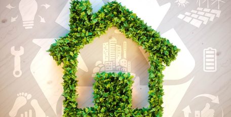Im Vordergrund steht der Umriss eines Hauses aus grünen Pflanzen, im Hintergrund weiße Symbole für Kreislaufwirtschaft, CO2-Fußabdruck, Solarenergie sowie eine geladene Batterie und ein Schraubenschlüssel, um das Konzept des umweltfreundlichen Bauens und Wohnen zu verdeutlichen.