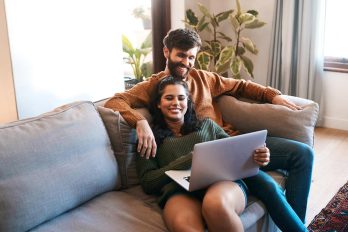 Ein Paar sitzt auf einem Sofa und lächelt, während sie einen Laptop betrachten. Die Frau sitzt vor dem Mann, der seine Arme um sie gelegt hat.