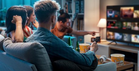 Vier Personen sitzen auf einem Sofa und schauen fern, mit Snacks und Getränken im Vordergrund.