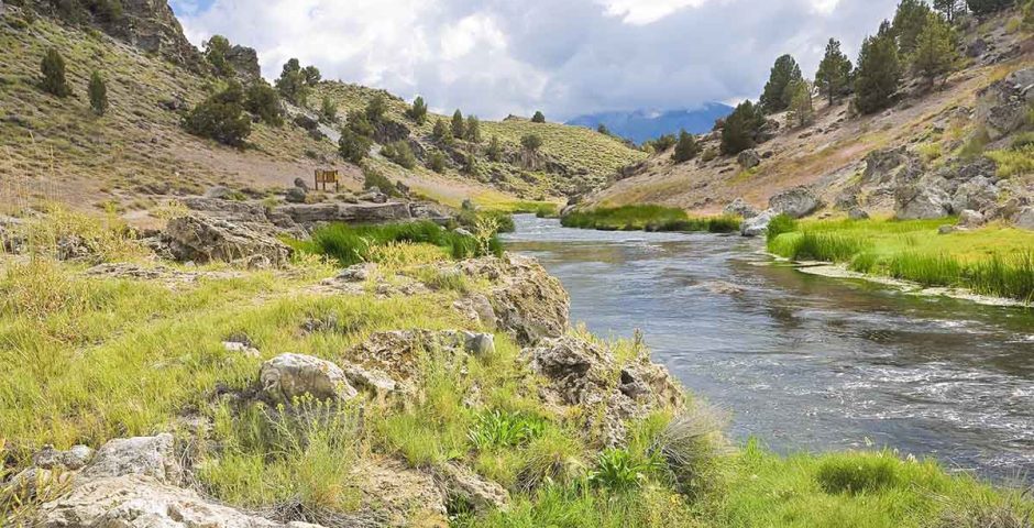 Ein malerischer Fluss fließt durch eine hügelige Landschaft mit grünen Wiesen und felsigen Ufern. Im Hintergrund sind Bäume und ein bewölkter Himmel zu sehen. Die Szenerie strahlt eine ruhige und friedliche Atmosphäre aus.