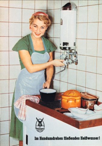 Eine Frau zapft in den 60er Jahren aus einem Kochendwassergerät von Vaillant Heißwasser für das Geschirr im Küchenspülbecken.