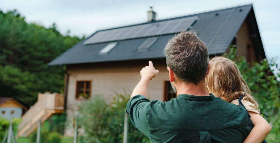 Rückansicht eines Vaters, der seine Tochter in den Armen hält und auf ein Haus mit installierten PV-Modulen zeigt