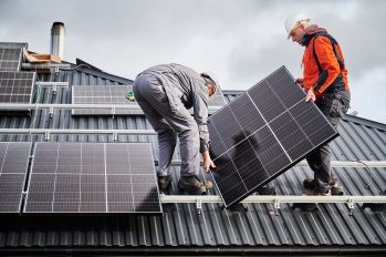 Zwei Techniker, die ein Photovoltaik-Modul tragen, während sie eine PV-Anlage auf dem Dach eines Hauses installieren