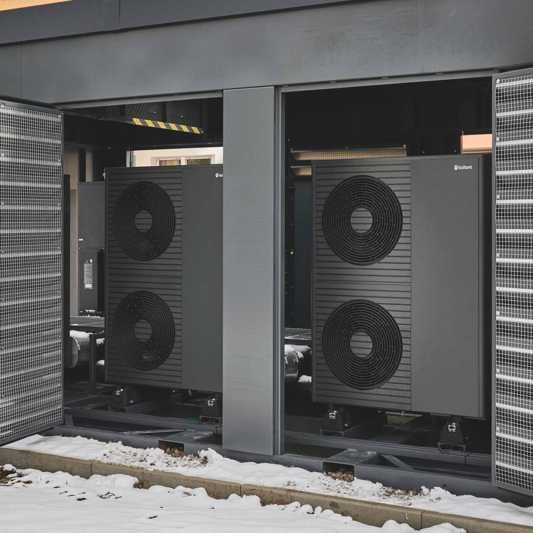 Außen aufgestellter Technikcontainer der Energy Unit mit geöffneten Türen, in dem zwei Luft/Wasser-Wärmepumpen von Vaillant mit jeweils zwei Ventilatoren vor einem Wohngebäude installiert sind.