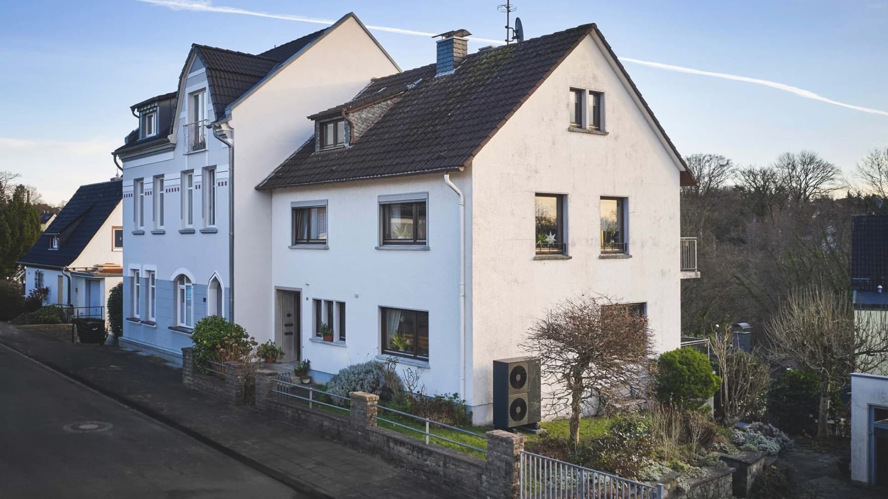 Freistehendes Einfamilienhaus mit hellem Fassadenanstrich und dunklem Dach, umgeben von einem gepflegten Garten mit Rasenflächen. Vor dem Haus befindet sich eine gepflasterte Einfahrt mit geparktem Fahrzeug, im Hintergrund sind weitere Wohnhäuser und eine ländliche Umgebung zu sehen
