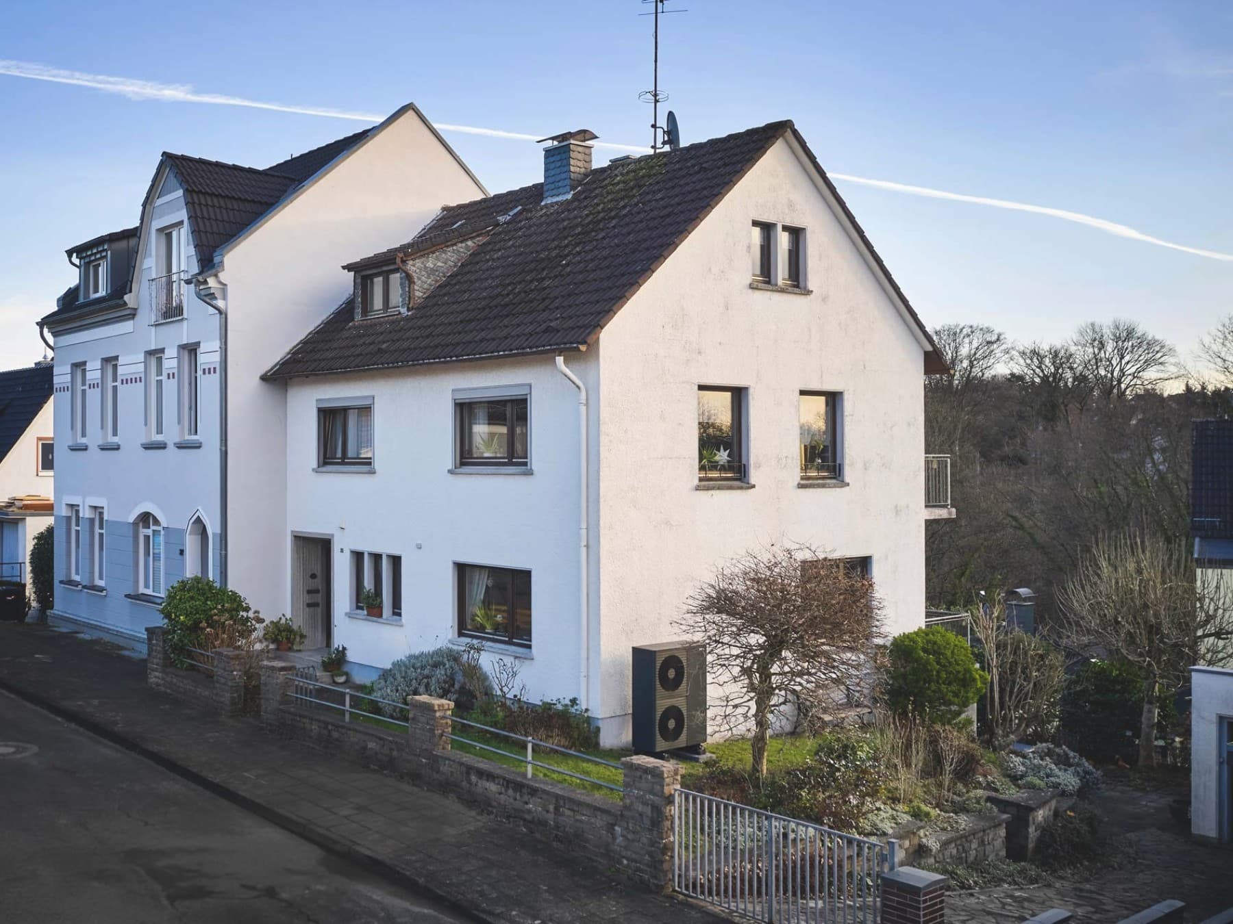 Zweistöckiges, weiß verputzte Haushälfte mit dunklem Dach, gelegen an einer ruhigen Wohnstraße. Vor dem Haus befindet sich ein kleiner, gepflegter Vorgarten mit Rasenfläche, niedriger Steinmauer und Metallzaun samt Gartentor. Rechts an der Hausfassade steht die Wärmepumpe aroTHERM plus von Vaillant. Seitlich führt eine gepflasterte Einfahrt zu einer Garage im Hintergrund. Angrenzend stehen weitere Wohnhäuser, im Hintergrund sind kahle Bäume zu sehen. Das Haus ist in warmes Morgenlicht getaucht.