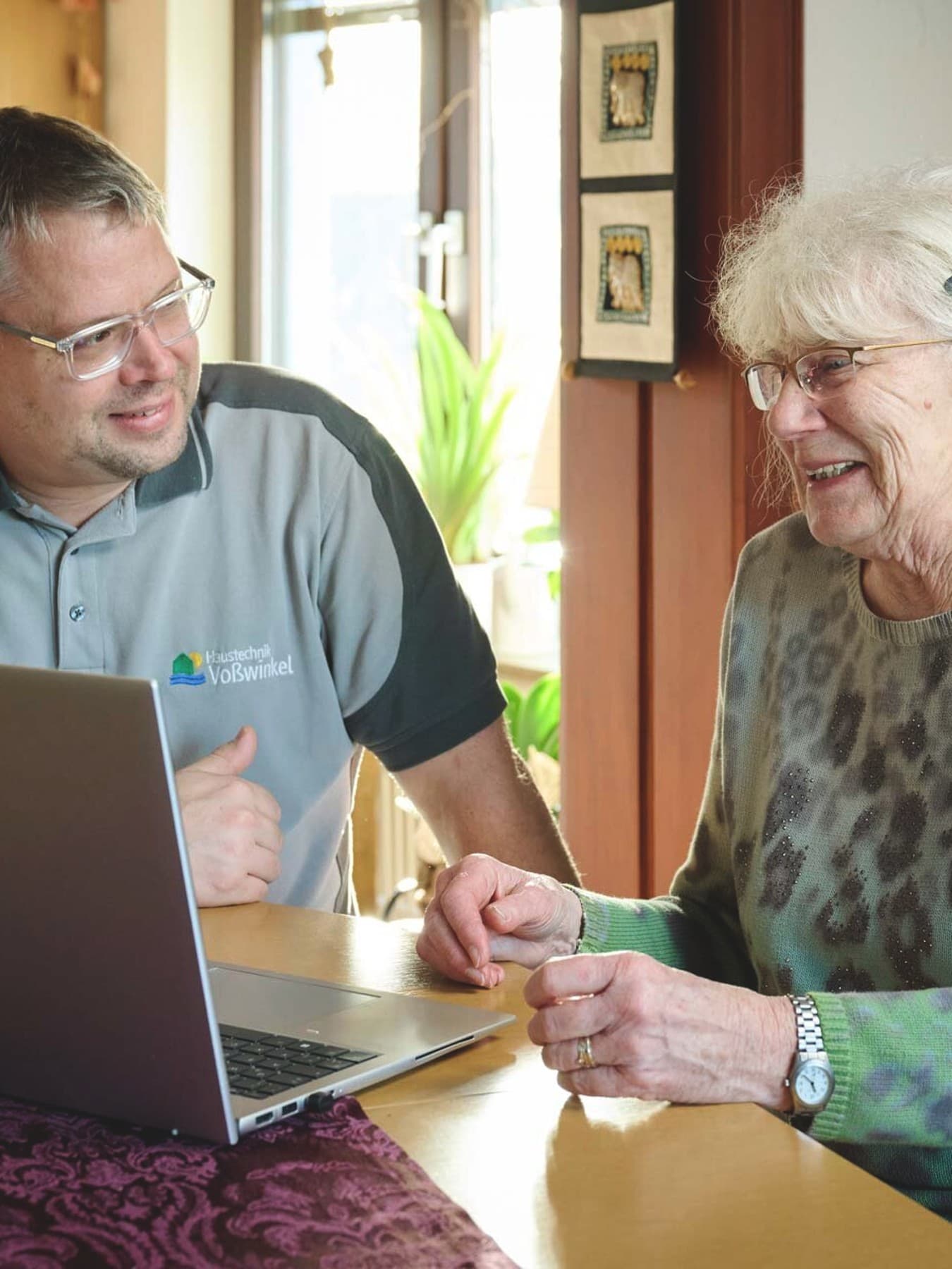 : Ein Mann und eine Frau sitzen gemeinsam an einem Esstisch in einem hellen Wohnraum. Vor ihnen steht ein aufgeklappter Laptop. Der Mann zeigt auf den Bildschirm und spricht, während die Frau lächelnd zuhört. Im Hintergrund sind Pflanzen und Wohnmöbel zu sehen, warmes Tageslicht fällt durch ein Fenster
