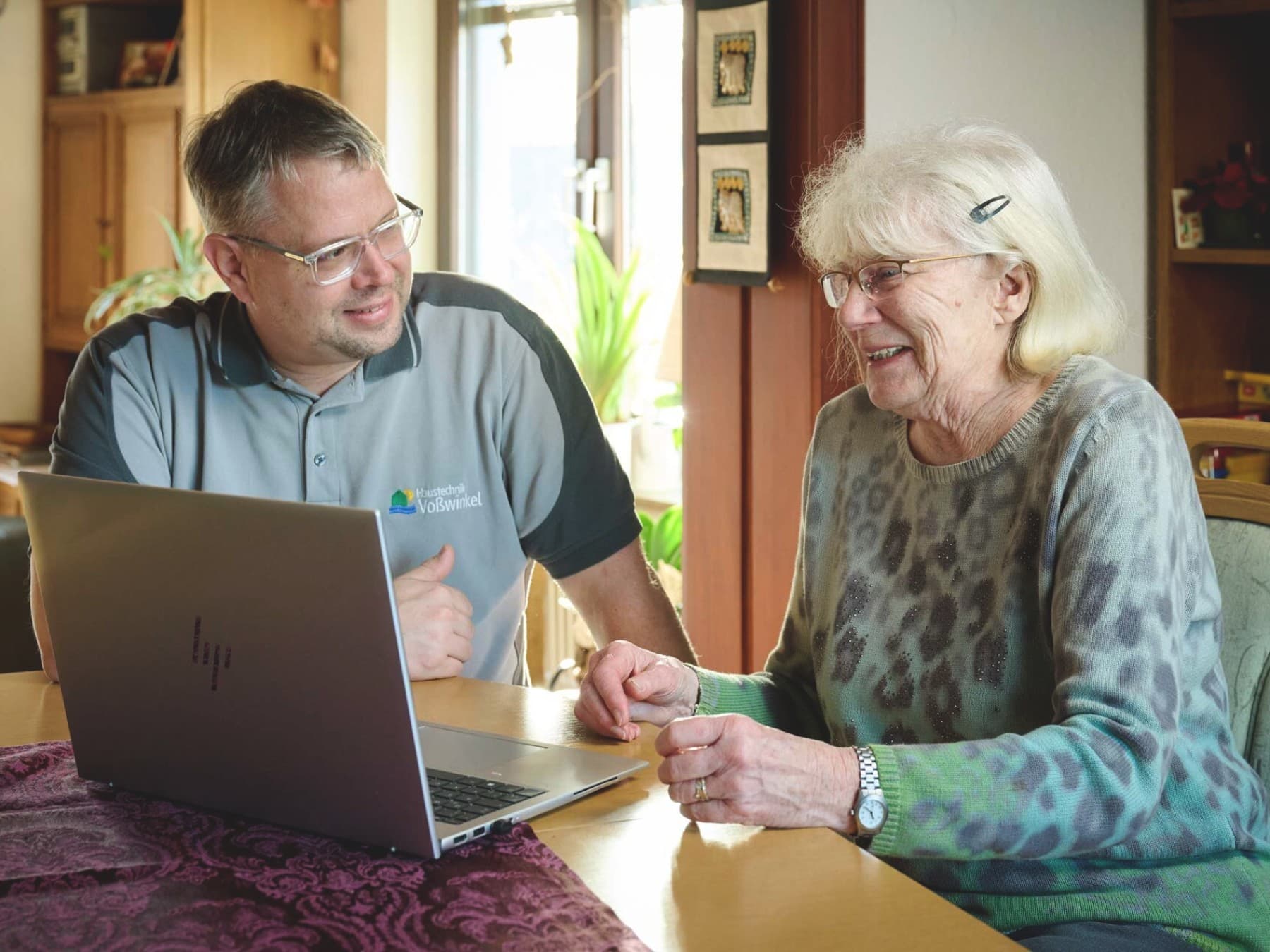 : Ein Mann und eine Frau sitzen gemeinsam an einem Esstisch in einem hellen Wohnraum. Vor ihnen steht ein aufgeklappter Laptop. Der Mann zeigt auf den Bildschirm und spricht, während die Frau lächelnd zuhört. Im Hintergrund sind Pflanzen und Wohnmöbel zu sehen, warmes Tageslicht fällt durch ein Fenster