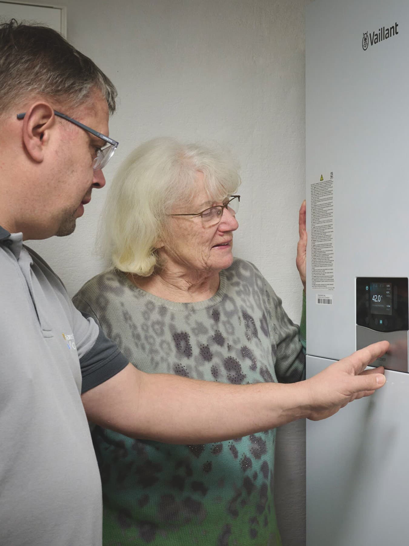Ein Mann und eine Frau stehen vor der Inneneinheit einer Wärmepumpe von Vaillant. Der Mann zeigt auf ein integriertes Display an der Front des Geräts, während die Frau aufmerksam hinsieht. Beide wirken im Gespräch.