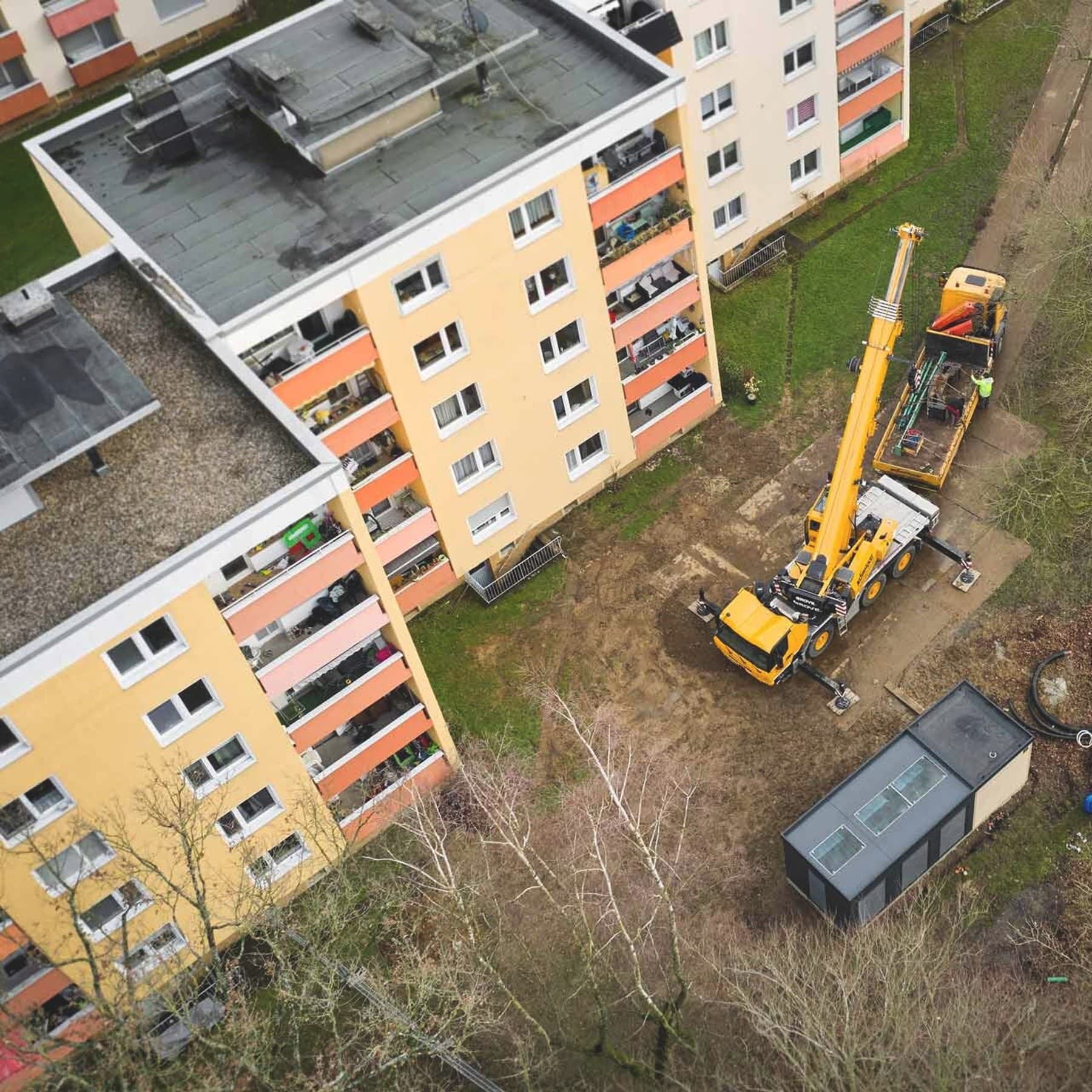 Luftaufnahme eines Krans beim Aufstellen eines Technikcontainers der Energy Unit neben zwei Mehrfamilienhäusern.