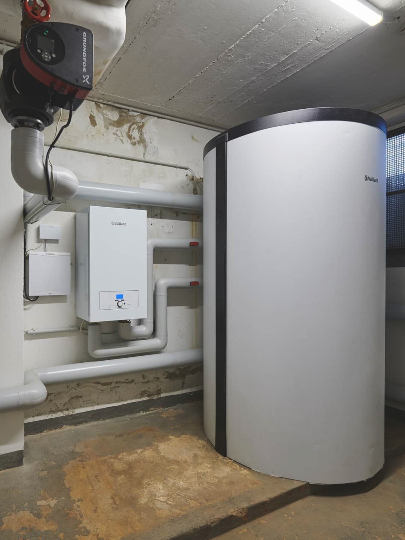 Heizungsraum im Keller mit an der Wand montierten Rohrleitungen. Ein großer 800 Liter Pufferspeicher steht auf der rechten Seite und links daneben ein Elektrozusatzheizgerät an der Wand vom Typ eloBLOCK von Vaillant.